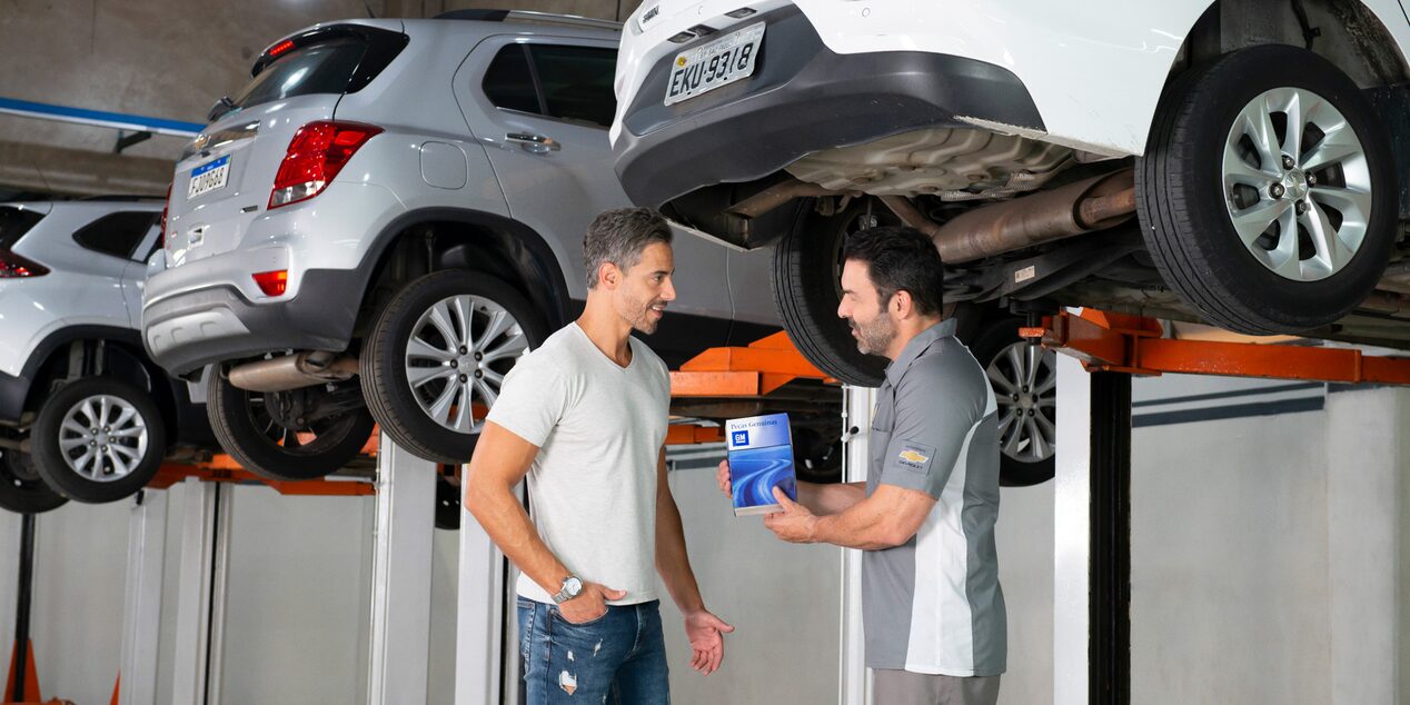 Imagen de un cliente mientras conversa con un mecánico en un taller Chevrolet, con varios vehículos elevados en elevadores hidráulicos para su revisión en el fondo.