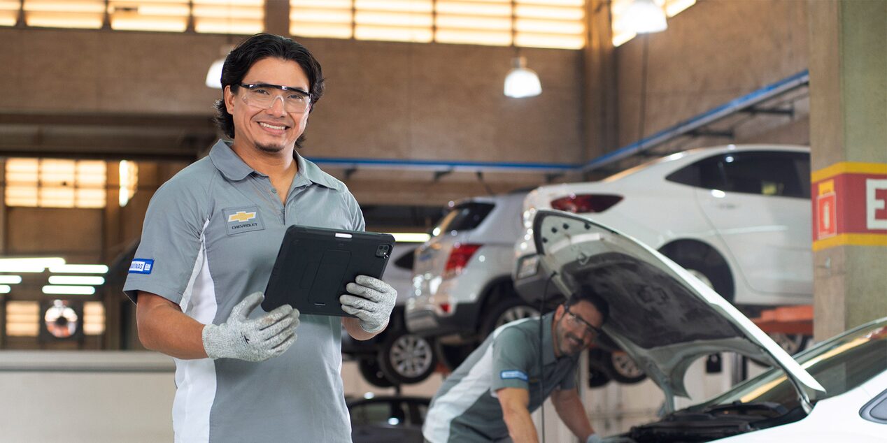 Imagen de un técnico de Chevrolet sonriendo mientras sostiene una tablet en un taller mecánico, con otro mecánico revisando el motor de un auto y varios vehículos elevados al fondo.